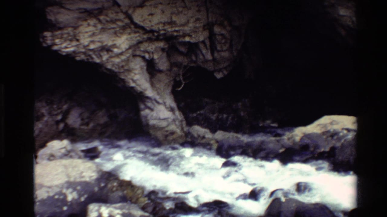 1980 Western U.S. 8mm frame of whitewater moving past rocks beneath a shadowed cave-like rock opening.