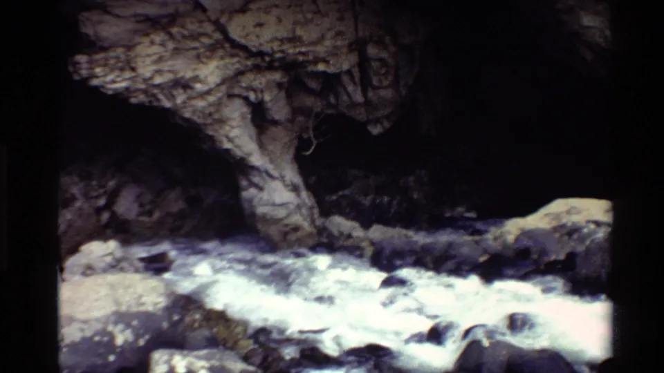1980 Western U.S. 8mm frame of whitewater moving past rocks beneath a shadowed cave-like rock opening.