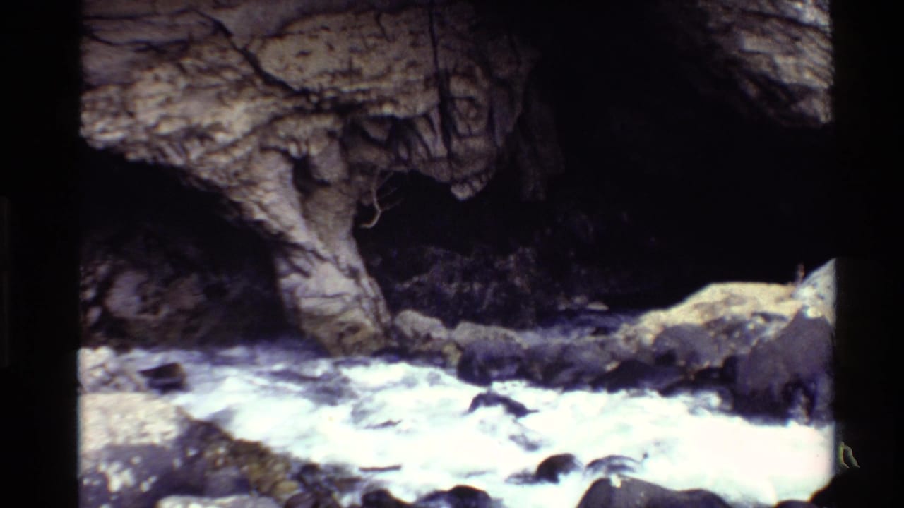 1980 Western U.S. 8mm frame showing a rocky stream flowing beside a shadowed cave or recess in the cliff.
