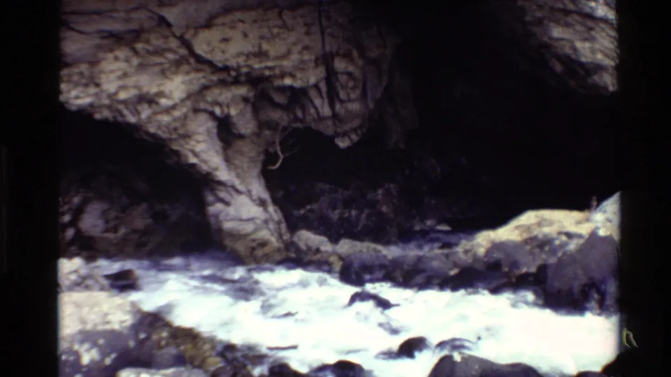 1980 Western U.S. 8mm frame showing a rocky stream flowing beside a shadowed cave or recess in the cliff.