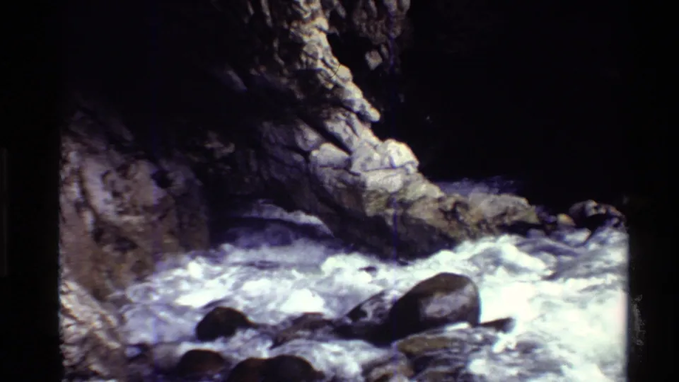 1980 Western U.S. 8mm frame of foaming water, large stream stones, and a rugged cave-like rock wall.