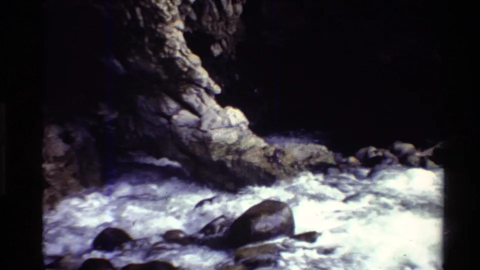 1980 Western U.S. 8mm frame with churning creek water, rounded stones, and a dark recess in rugged rock.