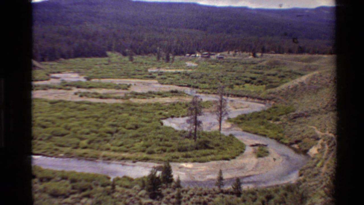 1980 Western United States 8mm frame showing a winding river, marshy green valley, scattered trees, and forested hills under a bright sky.