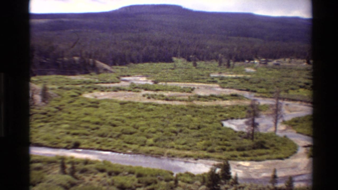 High-vantage 8mm view of a meandering river cutting through green marshland in the Western United States, with wooded hills on the horizon.