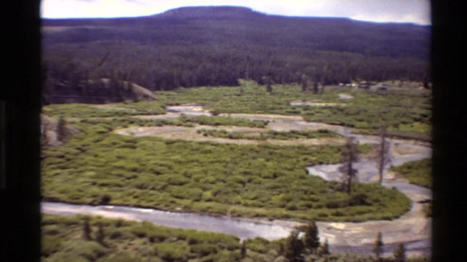 High-vantage 8mm view of a meandering river cutting through green marshland in the Western United States, with wooded hills on the horizon.