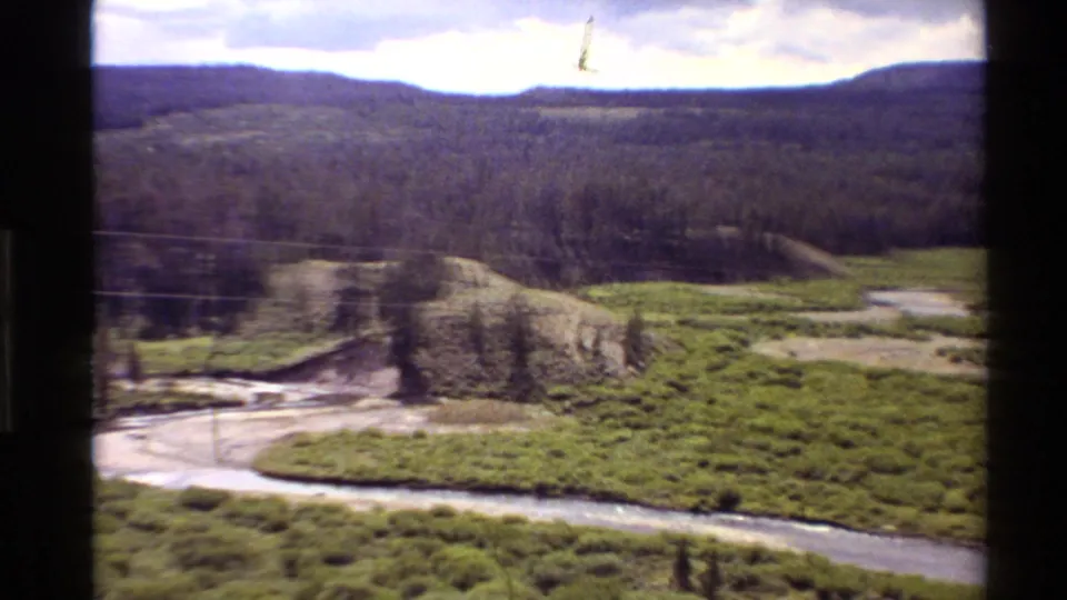 Soft 8mm frame of a river bend in a grassy Western United States valley, with scattered trees, pale banks, and distant wooded hills.