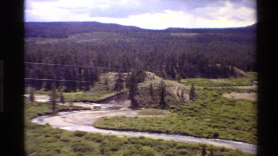 1980 Western U.S. 8mm view of a winding river crossing green meadow beneath pine forest, hills, and cloudy sky.