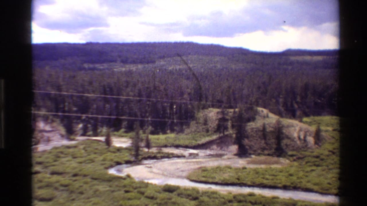 River bend, grassy meadow, pine forest, hills, and clouded sky.