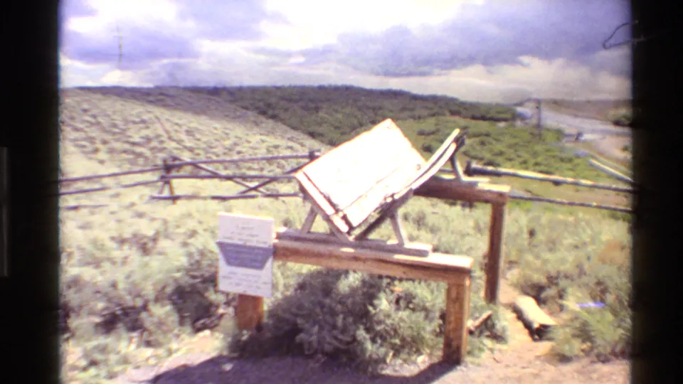 Visitor-overlook view with rustic wooden railings, a tilted sign, sage-like plants, and a river valley beyond in 1980 Western U.S. footage.