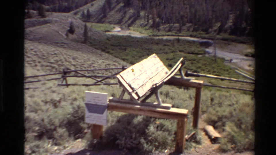 Rustic scenic overlook foreground with wooden fencing and sign structure, looking out to a winding river, meadow, and forested slope.