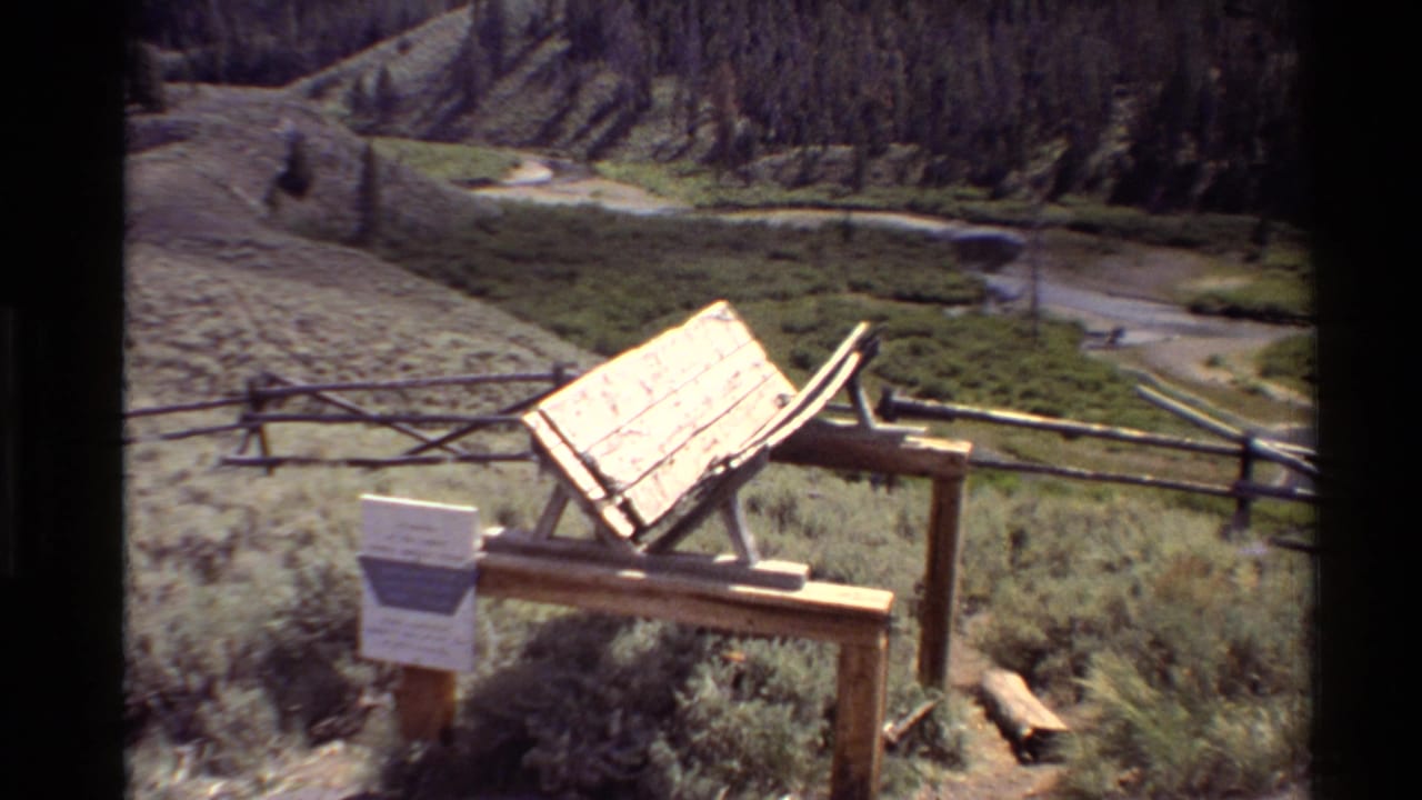 1980 Western USA home-movie frame showing a wooden information sign, rustic fence, green field, winding brook, and forested hillside.