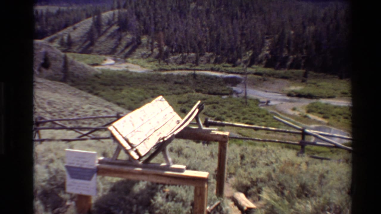 1980 Western USA frame with a sunlit wooden sign structure, small placard, split-rail fence, grassy meadow, brook, and wooded hill.