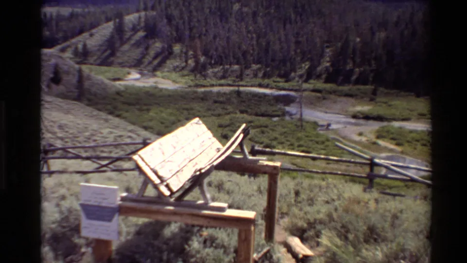1980 Western USA frame with a sunlit wooden sign structure, small placard, split-rail fence, grassy meadow, brook, and wooded hill.