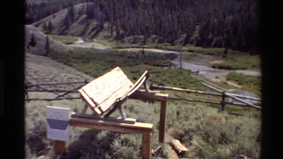 Western USA 1980 home-movie still showing a wooden sign stand, placard, rustic rail fence, open grass, winding water, and forested hillside.