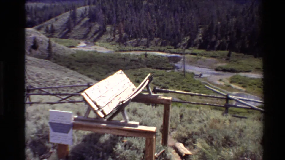1980 Western USA 8mm frame of a wooden interpretive sign and split-rail fence set above a green field, brook, and tree-lined slope.