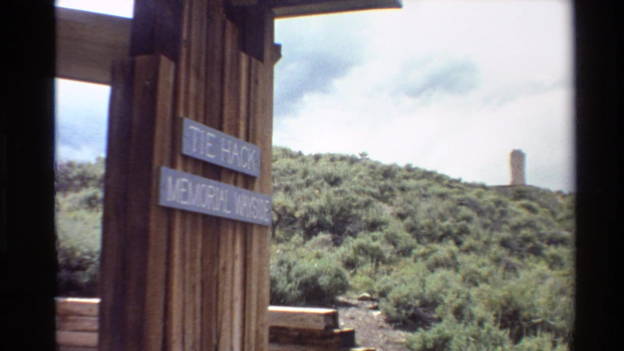 1980 Western U.S. 8mm frame showing a wooden Tie Hack Memorial Wayside sign, green shrubs, rolling hillside, and a distant stone monument.