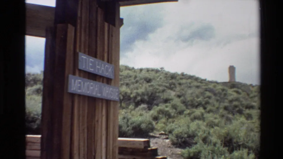 1980 8mm travel footage frame showing the memorial wayside sign closer in the foreground, with Western U.S. shrubs and a distant monument.
