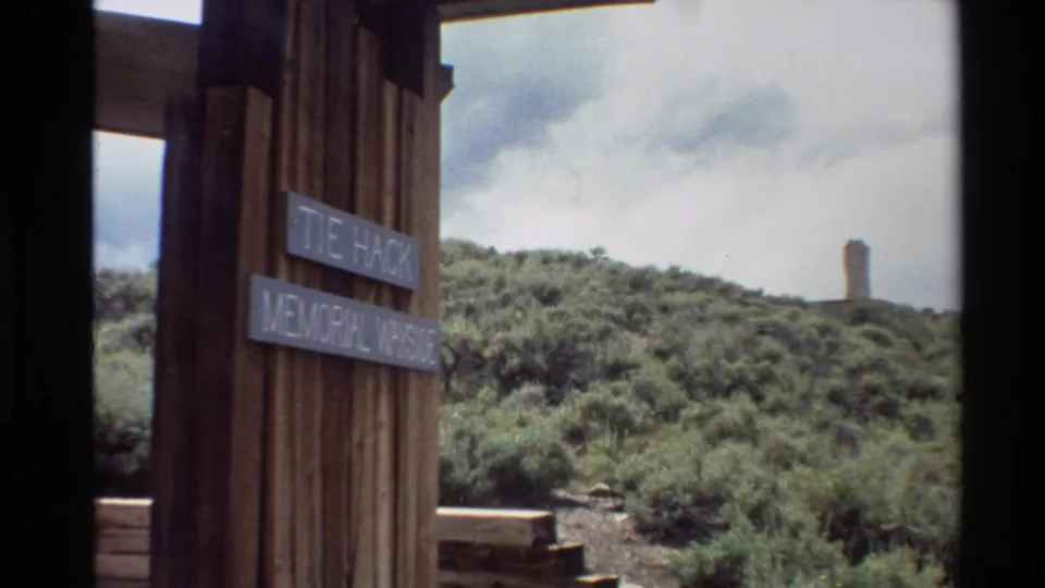 Western U.S. 1980 home-movie frame looking past a wooden wayside sign toward brushy hills and a solitary distant monument.