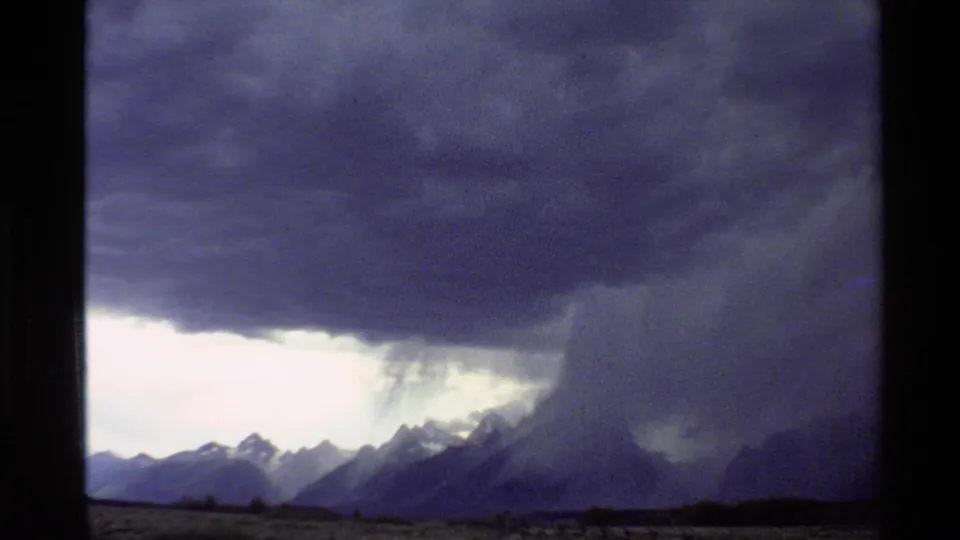 Grainy 8mm home-movie frame of a broad storm cloud over Western United States mountains, with rain visible near the ridge.
