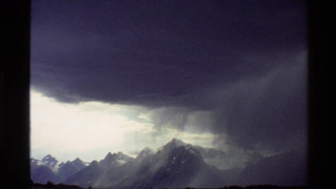 1980 Western USA 8mm frame showing storm clouds and rain shafts above distant snow-capped mountain ridges.