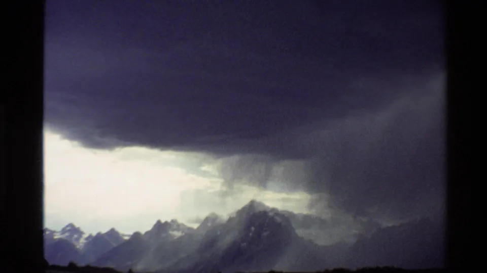 1980 Western USA 8mm frame showing storm clouds and rain shafts above distant snow-capped mountain ridges.