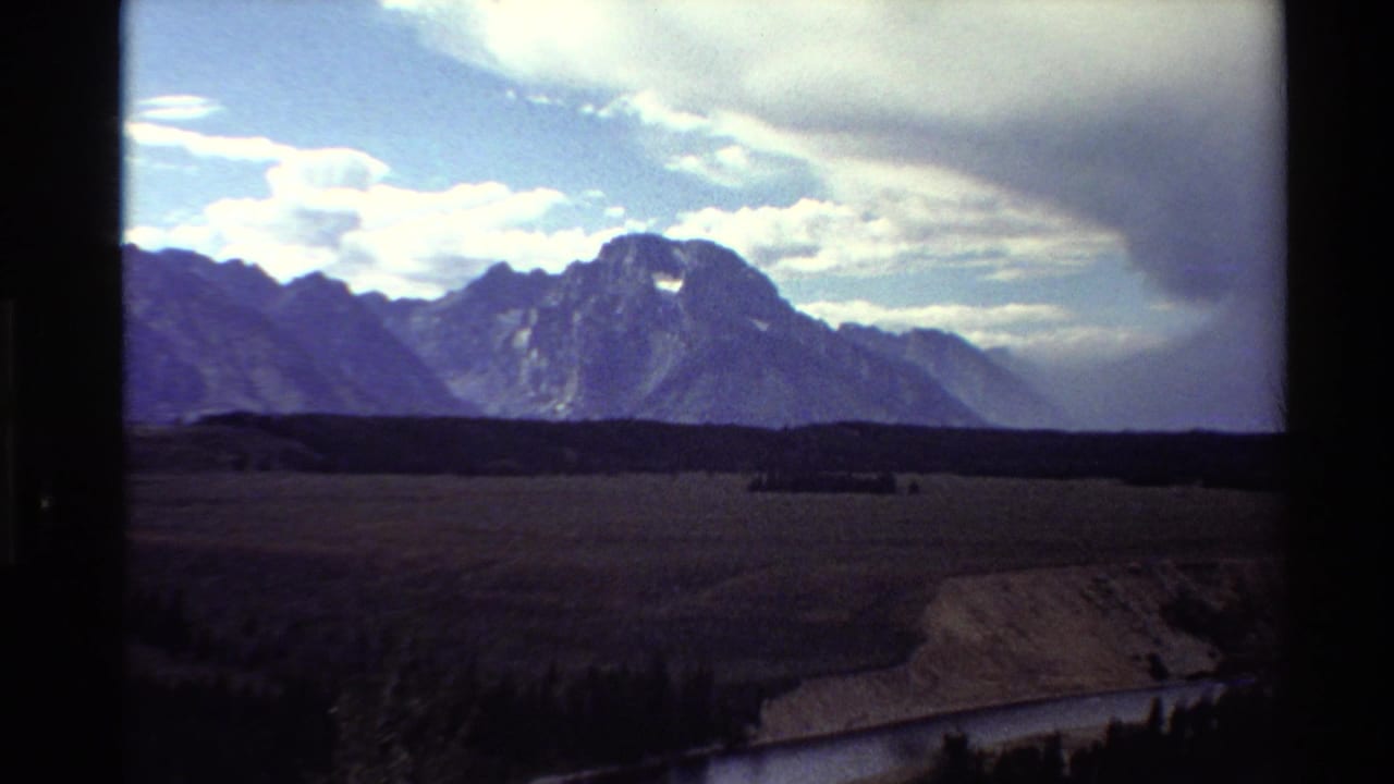 1980 Western USA 8mm frame showing a rugged mountain range, heavy cloud bank, open valley, dark tree line, and winding river.