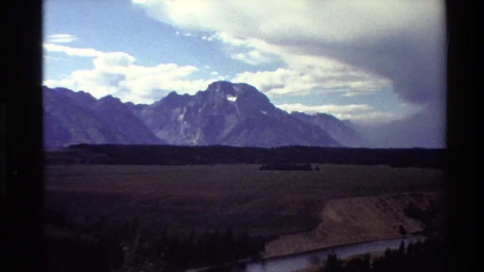 Western USA 1980 8mm frame showing a darkened valley foreground, distant mountain peaks, layered clouds, and a river bend near the lower edge.