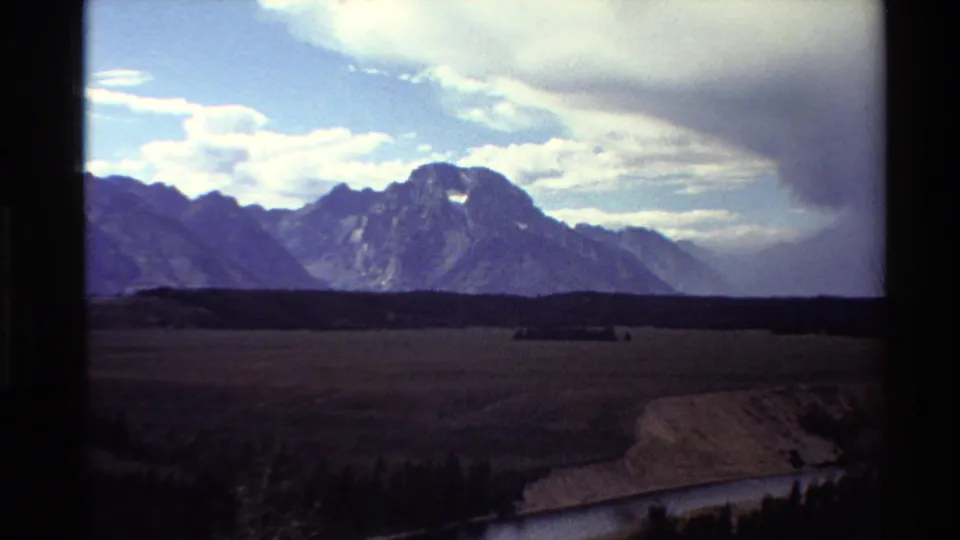 1980 Western USA 8mm frame with dark foreground trees, open valley, rugged mountains, river edge, and a heavy cloud bank overhead.