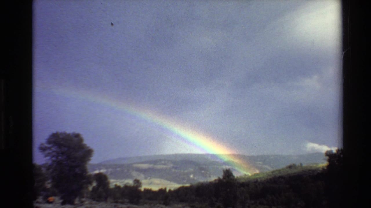 Rainbow over distant hills with trees and a dark foreground slope.