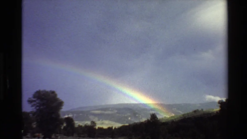 1980 Western U.S. 8mm view of a rainbow over rolling hills, with trees in silhouette and soft valley light below.