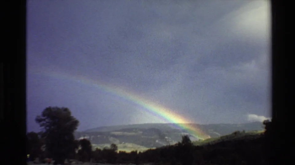 1980 Western U.S. 8mm frame with a low rainbow fading into a cloudy sky over trees, brush, and distant hills.