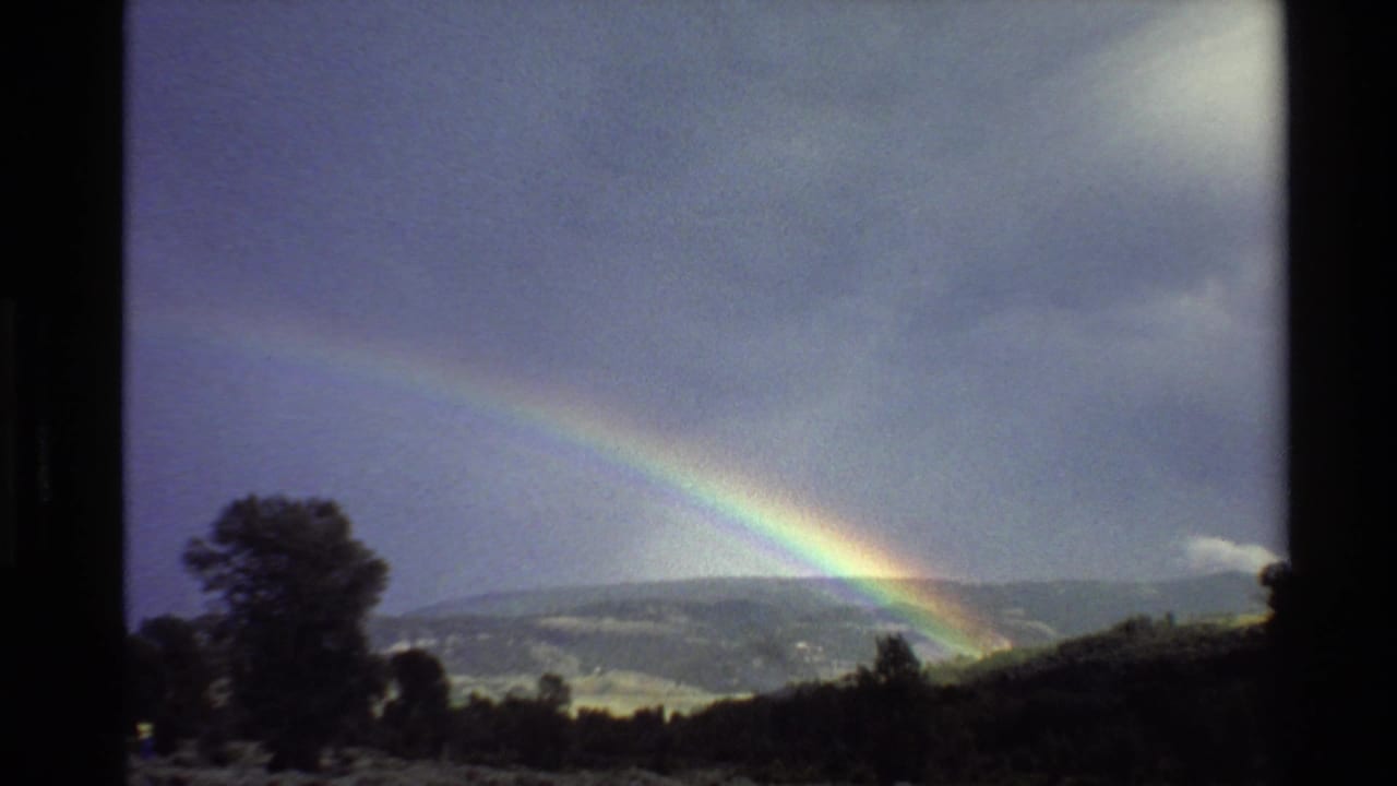 Double rainbow over hills beneath a dark cloudy sky, with trees in the foreground.