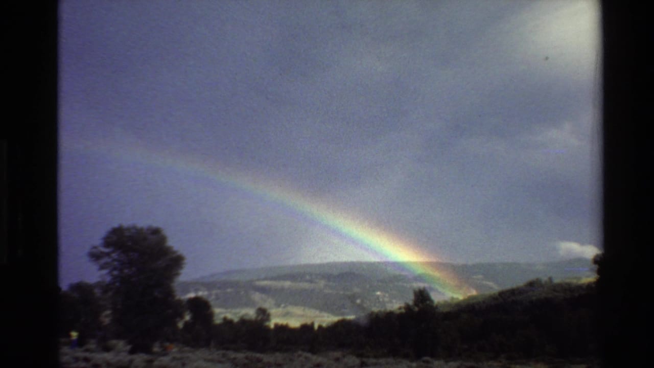 Rainbow over a hilly landscape with a dark tree silhouette and cloudy sky.
