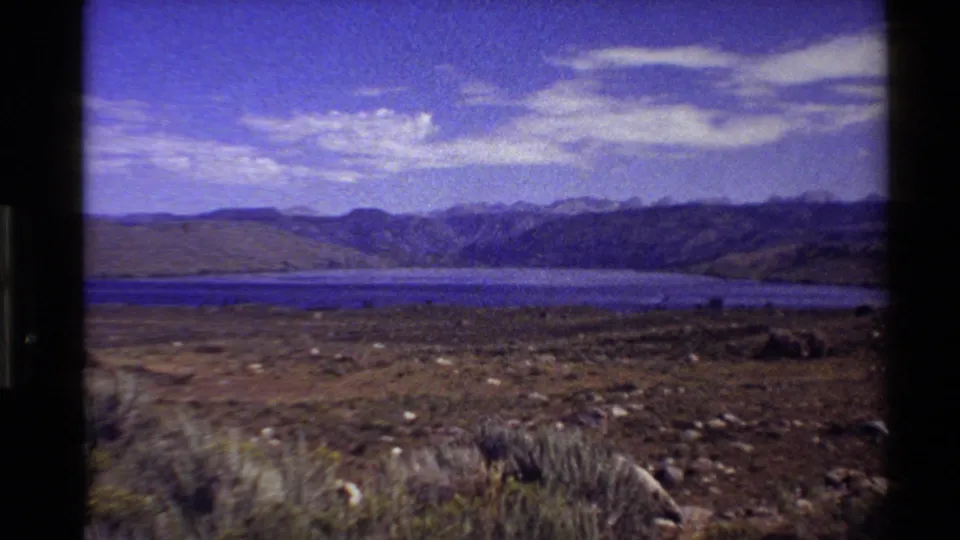 1980 Western USA 8mm frame showing a blue lake, arid foreground, distant mountains, and bright clouds in a wide landscape view.