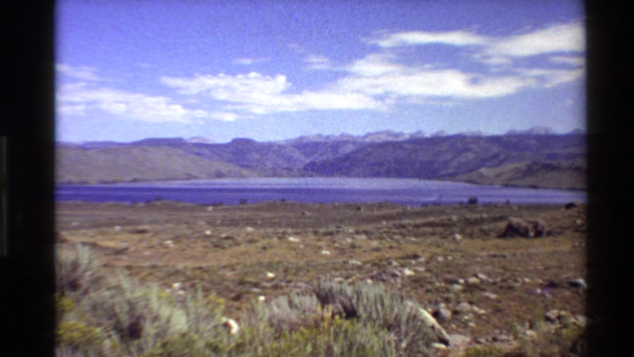 1980 Western U.S. 8mm frame showing dry foreground grasses, flat terrain, a blue lake, and distant mountains beneath pale clouds.