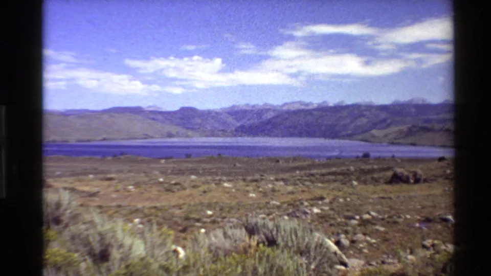 1980 Western U.S. 8mm frame showing dry foreground grasses, flat terrain, a blue lake, and distant mountains beneath pale clouds.