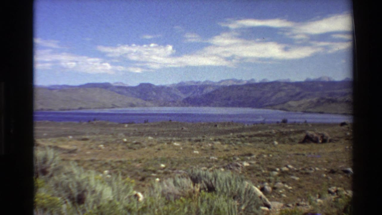 Western U.S. home-movie view of an arid lakeside landscape, with rocks and brush in front and mountain ridges in the distance.