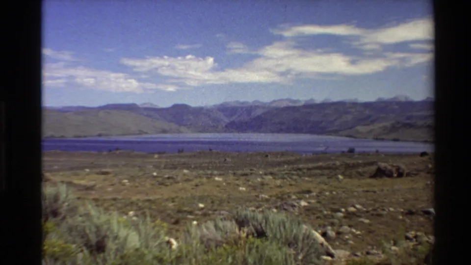 Aged 8mm frame from the Western United States showing dry foreground vegetation, open flatlands, a blue lake, and hazy mountains.