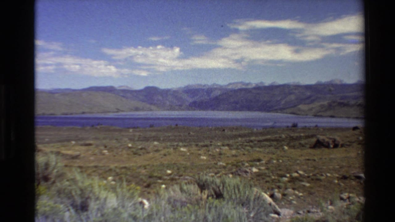 1980 Western U.S. 8mm frame showing a mountain lake, open scrubland, distant hills, and a partly cloudy sky.