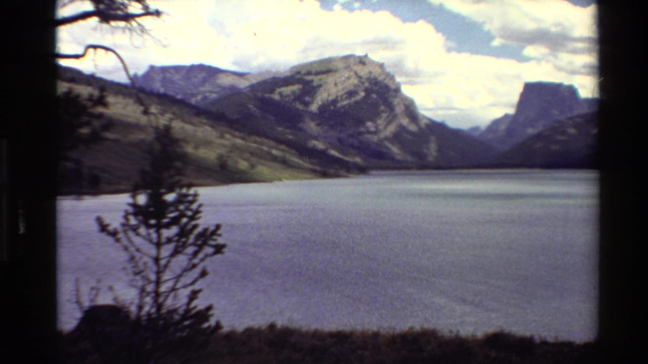 1980 Western U.S. 8mm view of a mountain lake bordered by shadowed trees and rugged peaks beneath clouds.