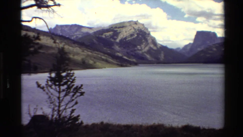1980 Western U.S. 8mm view of a mountain lake bordered by shadowed trees and rugged peaks beneath clouds.