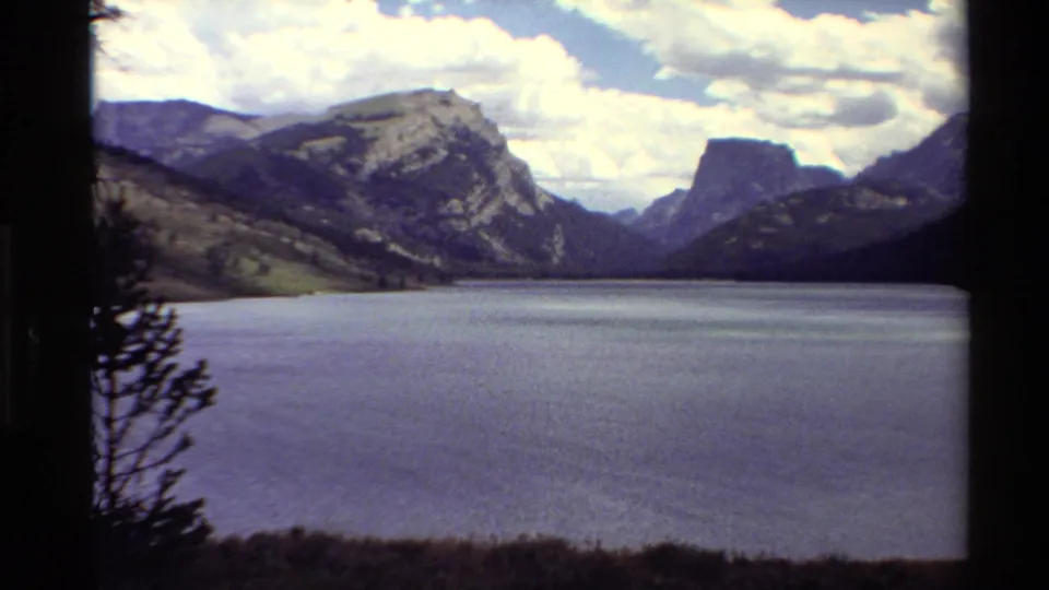 1980 Western U.S. 8mm frame with a wide mountain lake, silhouetted foreground tree, sloped hills, and bright clouds.
