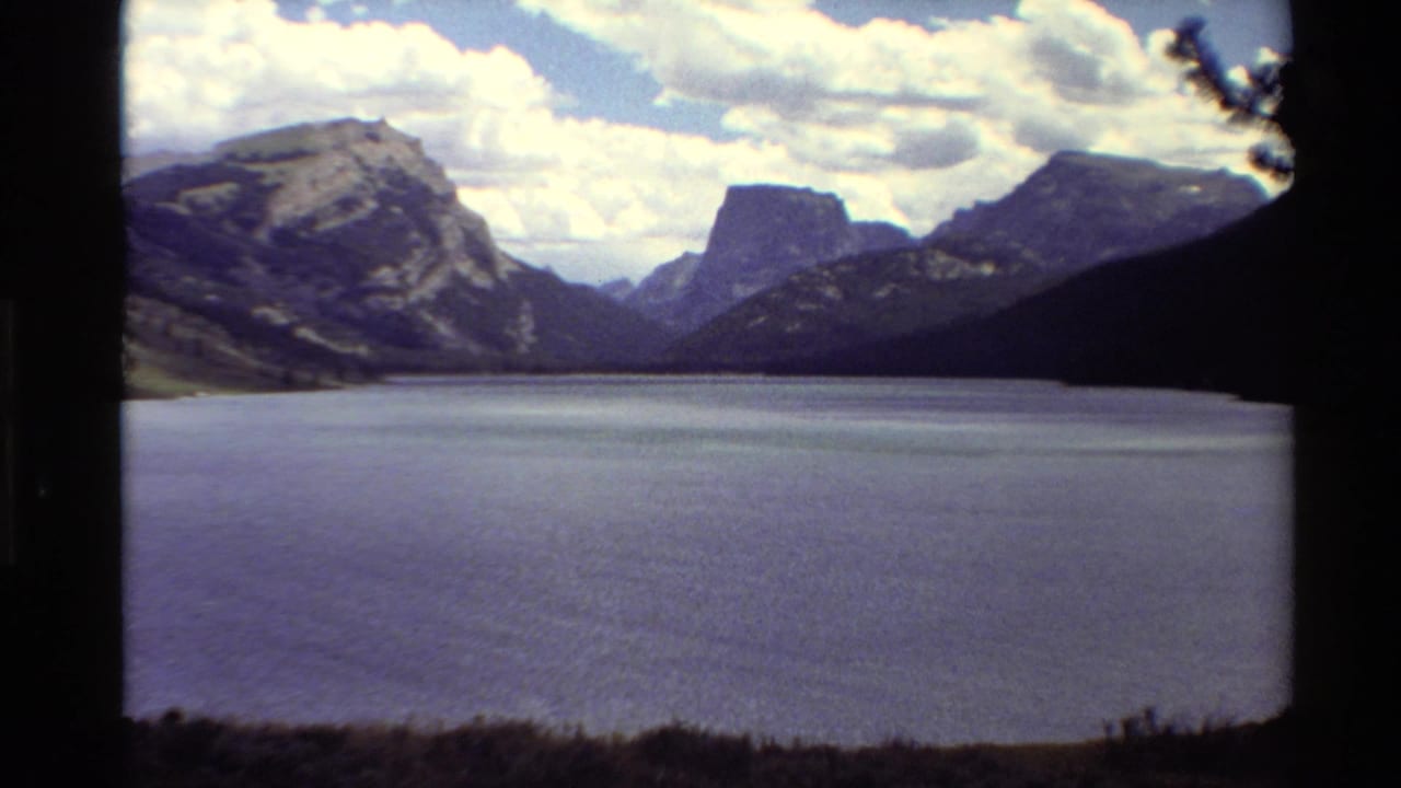 1980 Western U.S. 8mm frame of a broad alpine lake backed by rugged mountains, seen through a dark foreground edge.
