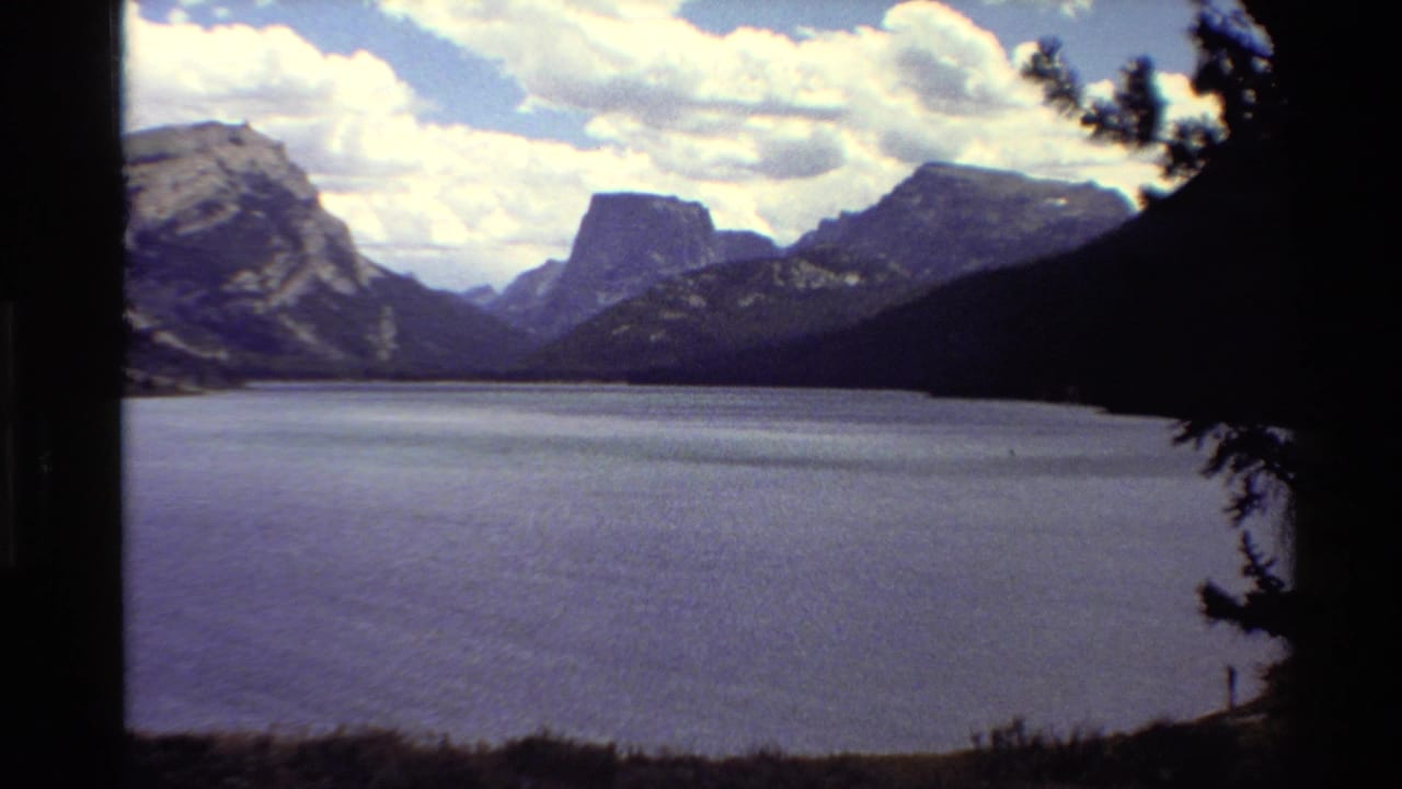 Wide 8mm view of a calm Western U.S. mountain lake with rugged peaks, clouds, and dark pine framing at the edge.