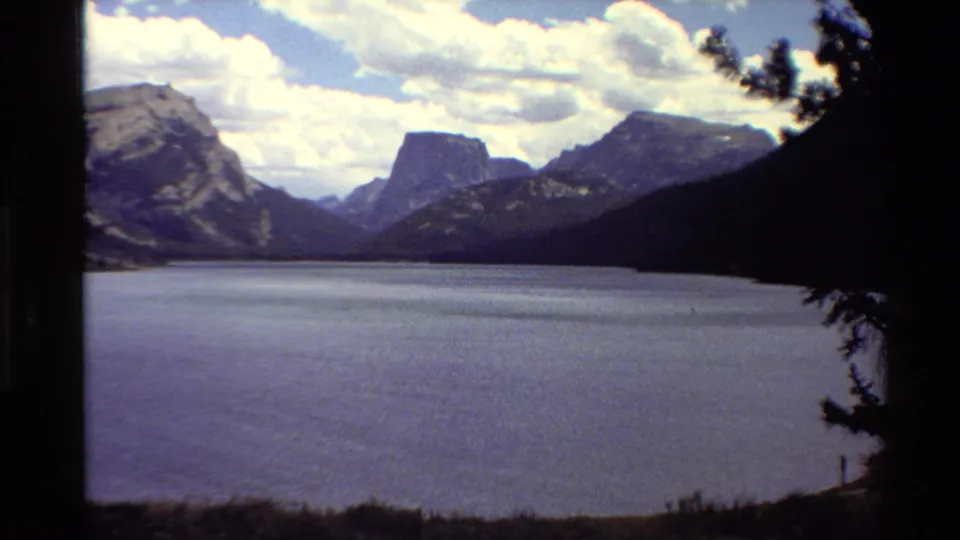 Wide 8mm view of a calm Western U.S. mountain lake with rugged peaks, clouds, and dark pine framing at the edge.