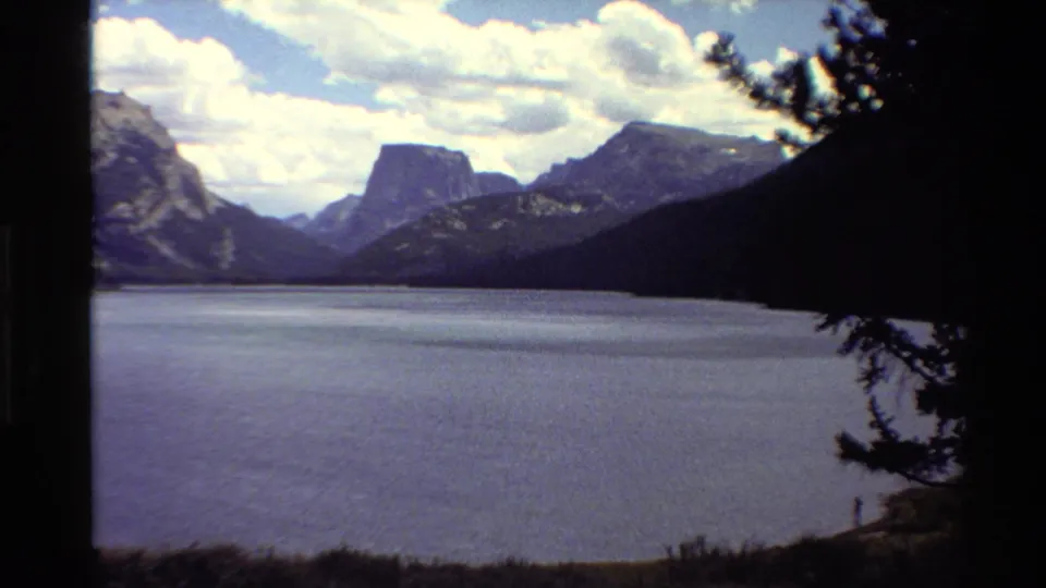 1980 home-movie frame of a broad lake below mountains, with pine branches and a tiny possible person near the shoreline.
