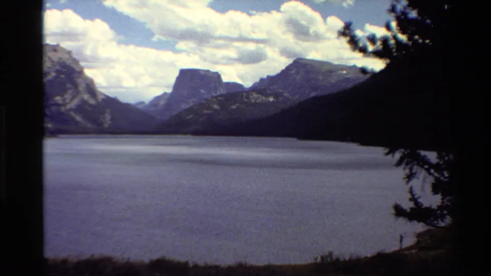 Soft 8mm frame of a Western U.S. mountain lake under clouds, with dark tree framing and shoreline vegetation in front.