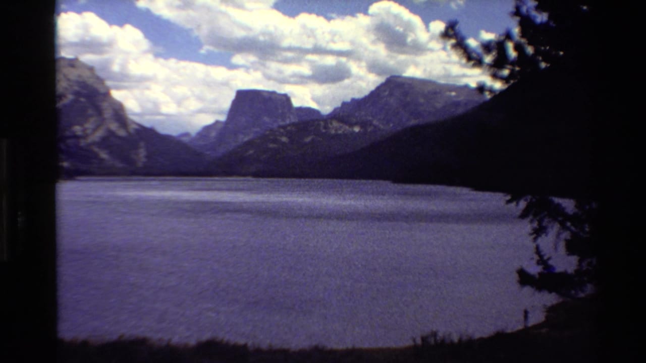 1980 Western USA 8mm frame showing a wide mountain lake, rugged peaks, bright clouds, and dark tree branches along the edge.