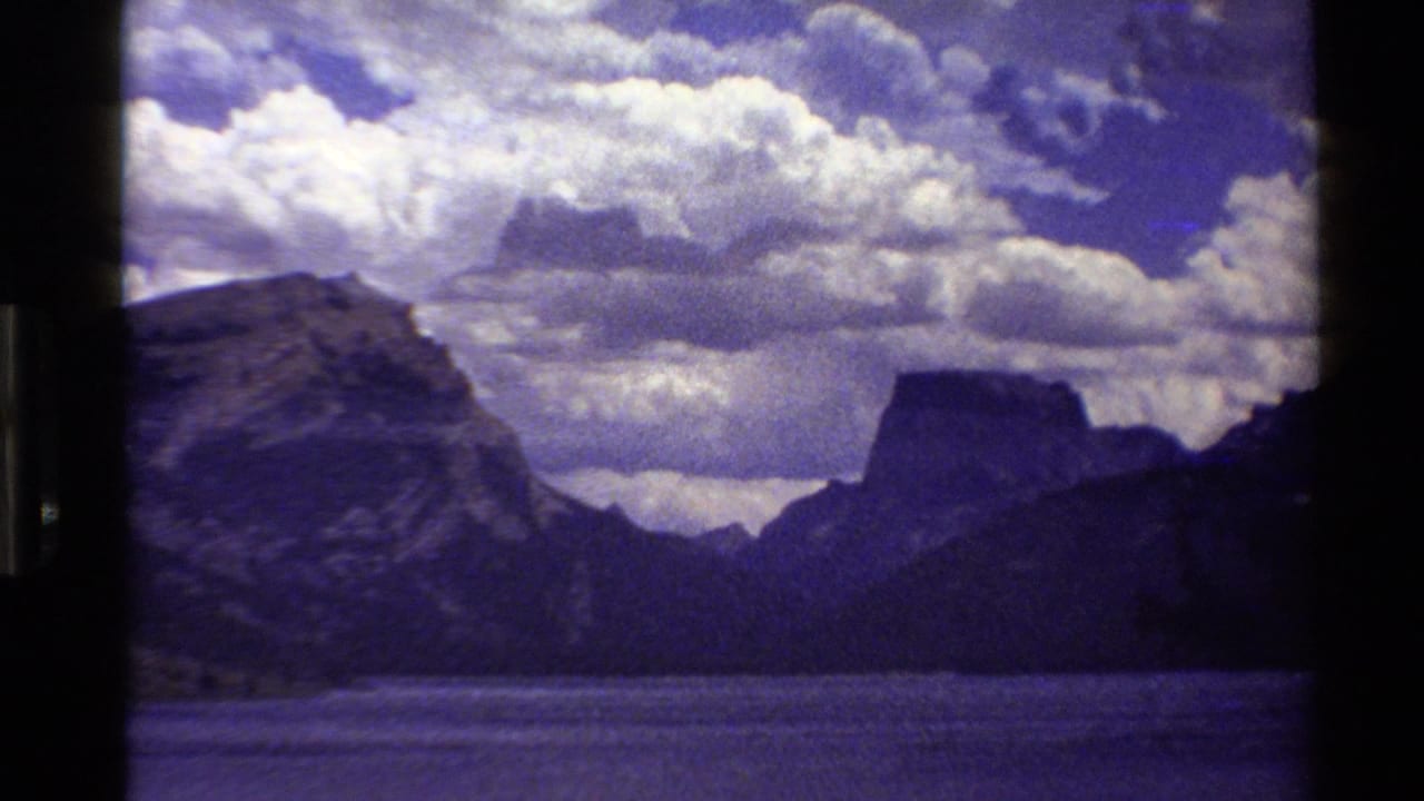1980 Western USA 8mm frame with rocky mountain slopes, a distant flat-topped peak, a lake surface, and large white clouds.