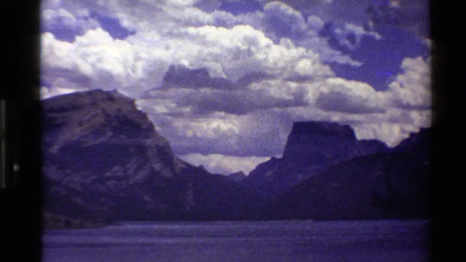 1980 Western USA 8mm frame with rocky mountain slopes, a distant flat-topped peak, a lake surface, and large white clouds.